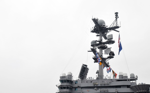 The image shows the flight deck of USS Abraham Lincoln (CVN-72), a Nimitz-class aircraft carrier, during what appears to be a formal naval ceremony or muster. A large formation of sailors dressed in white uniforms is assembled on the flight deck facing the island superstructure. Multiple aircraft and support equipment are visible on the deck.