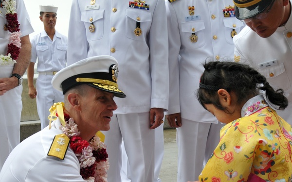A senior naval officer in white dress uniform, wearing a floral lei and gold shoulder boards, kneels down to interact with a young girl dressed in a traditional Korean hanbok (yellow jeogori top and pink skirt). The scene appears to be a formal ceremonial welcome or greeting event, with multiple naval officers in white dress uniforms standing in the background. The setting suggests an official diplomatic or military visit, likely in South Korea.