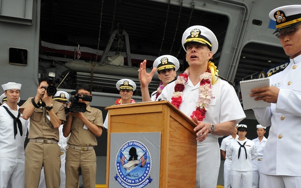 A U.S. Navy officer wearing a floral lei is delivering a speech at a podium bearing the USS George Washington (CVN-73) emblem, suggesting a ceremonial arrival or departure event. The scene takes place on a pier or flight deck apron with a large naval vessel visible in the background. Multiple sailors in dress white and working uniforms are present, along with a videographer documenting the event.