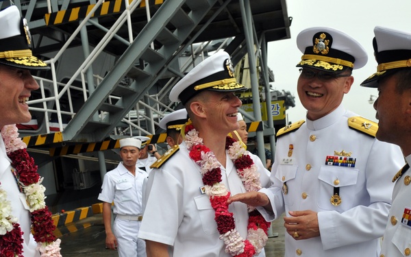 A group of naval officers in white dress uniforms are gathered at what appears to be a port or naval facility. Two officers are wearing floral leis, suggesting a ceremonial welcome or arrival event. The officers appear to be engaged in friendly conversation, indicating a diplomatic or official military exchange.