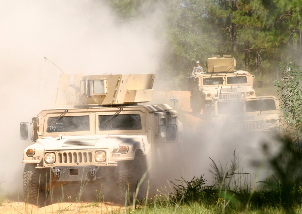 A convoy of military HMMWV (Humvee) vehicles is traveling along a dusty dirt trail through a forested area. The vehicles are kicking up significant dust clouds as they move, suggesting dry conditions and rapid movement. At least one soldier is visible manning a turret position on one of the rear vehicles.