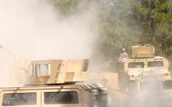 A convoy of military HMMWV (Humvee) vehicles is traveling along a dusty dirt trail through a forested area. The vehicles are kicking up significant dust clouds as they move, suggesting dry conditions and rapid movement. At least one soldier is visible manning a turret position on one of the rear vehicles.