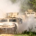 A convoy of military HMMWV (Humvee) vehicles is traveling along a dusty dirt trail through a forested area. The vehicles are kicking up significant dust clouds as they move, suggesting dry conditions and rapid movement. At least one soldier is visible manning a turret position on one of the rear vehicles.