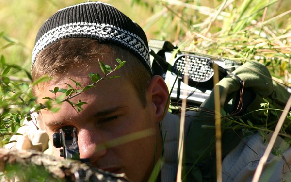 A young male is lying prone in dense vegetation, aiming a firearm with a scope through low brush. He is wearing a black and white knit cap and olive/tan clothing, with a backpack visible on his back. The setting appears to be an outdoor field environment with dry grass and green shrubs.