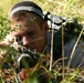 A young male is lying prone in dense vegetation, aiming a firearm with a scope through low brush. He is wearing a black and white knit cap and olive/tan clothing, with a backpack visible on his back. The setting appears to be an outdoor field environment with dry grass and green shrubs.