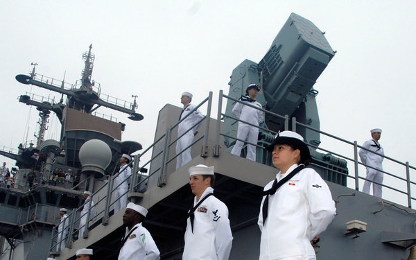A group of U.S. Navy sailors in dress white uniforms are manning the rails of a gray naval warship, likely during a formal arrival or departure ceremony. Multiple sailors are positioned at various levels of the vessel, from the main deck up through elevated platforms and superstructure. The ship features visible weapons systems, radar equipment, and communication masts.