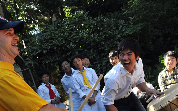 A Western adult male wearing a yellow shirt and blue cap is playing traditional barrel drums alongside an enthusiastic young Asian male, surrounded by a group of smiling children and teenagers. The setting appears to be an outdoor cultural exchange activity, likely in an East Asian country. The atmosphere is joyful and energetic, with participants laughing and engaging with the music.