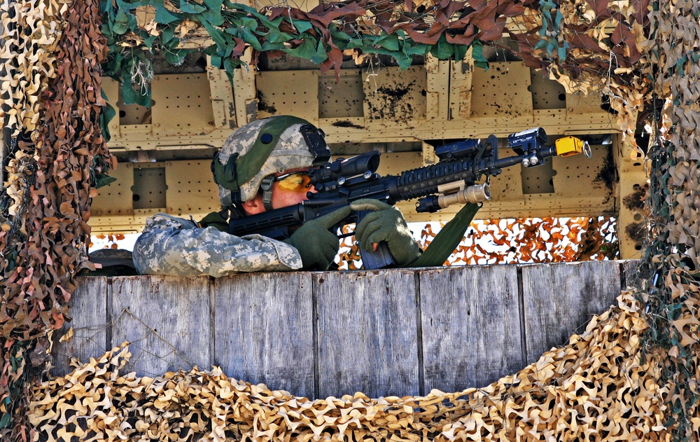 A soldier in U.S. Army ACU (Army Combat Uniform) digital camouflage is positioned at a fortified observation or firing position, aiming a rifle through an opening framed by camouflage netting. The position appears to be a constructed wooden platform or bunker covered with multi-terrain camouflage netting in tan, brown, and green patterns. The soldier is in a prone or low-crouch firing stance, actively sighting down the weapon's optic.