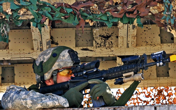 A soldier in U.S. Army ACU (Army Combat Uniform) digital camouflage is positioned at a fortified observation or firing position, aiming a rifle through an opening framed by camouflage netting. The position appears to be a constructed wooden platform or bunker covered with multi-terrain camouflage netting in tan, brown, and green patterns. The soldier is in a prone or low-crouch firing stance, actively sighting down the weapon's optic.