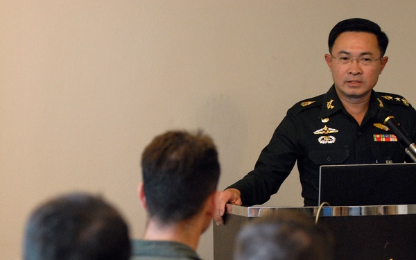 A senior military officer in a dark green uniform with medals and insignia stands at a podium addressing an audience. The podium bears a gold emblem featuring wings and a sunburst design consistent with Southeast Asian military insignia. Several audience members in military flight suits are visible in the foreground, seated with their backs to the camera.