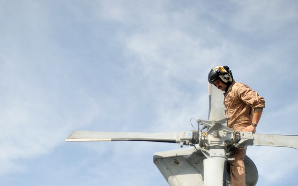 A technician or military maintenance crew member is perched atop the tail rotor assembly of a military helicopter, performing an inspection or maintenance task. The photo is taken from a low angle looking upward, emphasizing the height and scale of the work being performed. The individual is wearing flight deck gear including a helmet with visor and tan/khaki uniform.