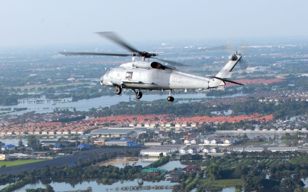 A U.S. Navy helicopter, marked with 'NAVY' on its fuselage and bearing a side number (615), flies over a heavily flooded landscape. The aerial view reveals extensive inundation of agricultural fields, residential neighborhoods, and roads. The scene is consistent with a disaster relief or reconnaissance mission over a flood-affected region.