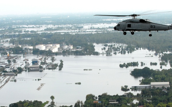 A military helicopter marked '613' conducts an aerial survey over a severely flooded urban and rural landscape. Extensive floodwaters have inundated residential neighborhoods, agricultural fields, and industrial areas as far as the eye can see. The image appears to document a disaster response or reconnaissance mission over a major flood event.