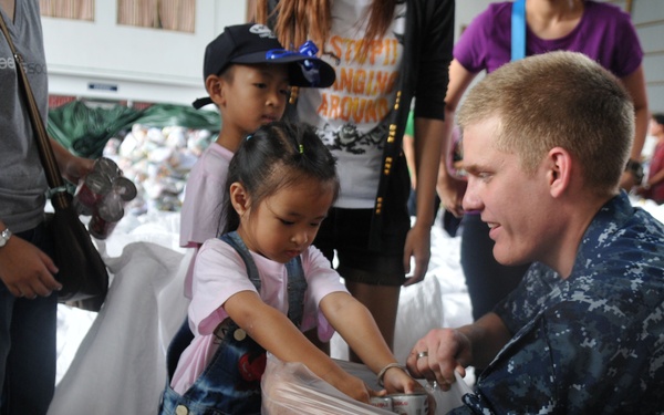 A U.S. Navy sailor in digital camouflage uniform crouches down to assist a young girl receiving supplies in what appears to be a humanitarian aid distribution event. The setting looks like a large indoor facility filled with bags of supplies and multiple civilians, including children and adults. The interaction suggests a disaster relief or community assistance operation.