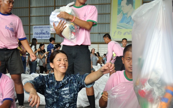 A U.S. Navy service member in digital camouflage uniform sits on the floor smiling while participating in what appears to be a humanitarian aid distribution operation alongside Thai volunteers wearing pink shirts. The setting appears to be a large indoor facility such as a warehouse or hangar where relief supplies are being assembled and packaged. The scene suggests a joint U.S.-Thai military humanitarian assistance or disaster relief effort.