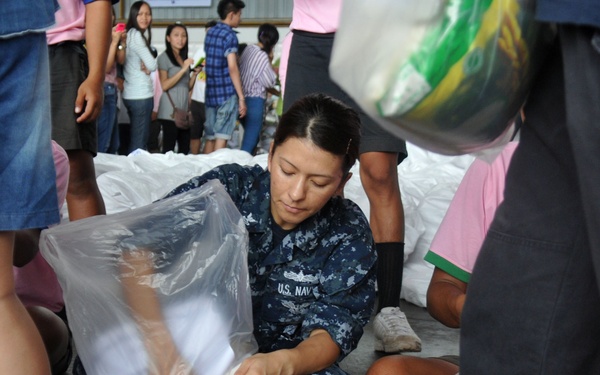 A U.S. Navy service member in digital camouflage uniform is kneeling on the ground packing or organizing a large clear plastic bag containing rice and other supplies. The scene appears to be a humanitarian aid or disaster relief distribution operation, with multiple civilians and other personnel visible in the background. Several bags of rice and other food supplies are visible throughout the scene.