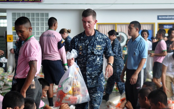 A U.S. Navy sailor, identified by name tape 'TAIT' and U.S. Navy digital camouflage uniform, distributes food supplies to a large group of young people wearing matching pink shirts. The setting appears to be an indoor facility, likely in Thailand based on the Thai script visible on signage and the pink uniforms. This appears to be a humanitarian aid or community outreach event involving U.S. military personnel and local youth.