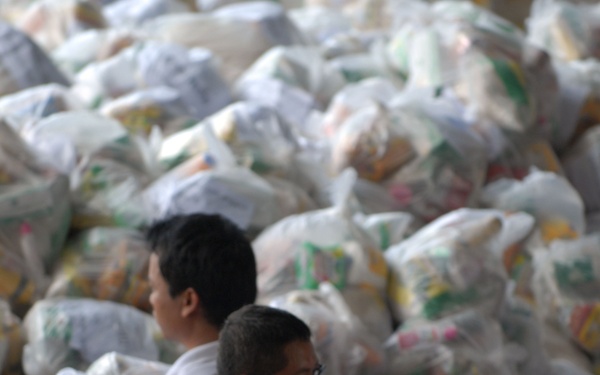 The image shows multiple individuals engaged in what appears to be a humanitarian aid or relief distribution operation. A person wearing a blue camouflage military uniform is prominently holding a white bag printed with Thai script reading 'กองทัพเรือ' (Royal Thai Navy). Behind them is a large pile of similarly packaged white supply bags, suggesting a significant stockpile of relief goods.