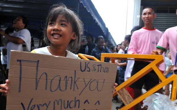 A young girl, likely Thai, holds a handwritten cardboard sign reading 'US &amp; Thai Thank you Very much :) We so Proud of you' in a crowded outdoor or semi-covered area. The scene appears to be a humanitarian or military aid event involving US and Thai personnel. The girl is smiling broadly, expressing gratitude on behalf of the local community.