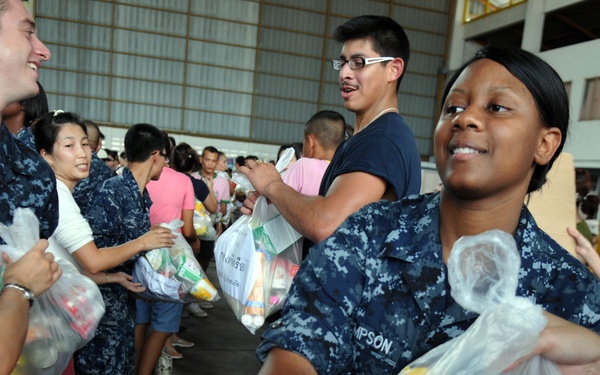 U.S. Navy personnel in digital camouflage uniforms are distributing bags of supplies or food items to civilians in what appears to be a humanitarian aid or community outreach operation. The event is taking place inside a large hangar or warehouse facility. Both military members and civilians of apparent Asian descent are participating in the distribution effort.