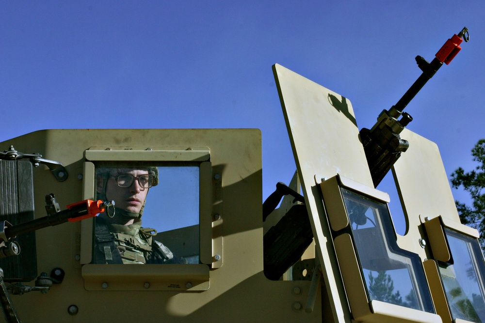 A soldier wearing glasses and combat gear is visible through the armored window port of a military vehicle, likely a HMMWV (Humvee) or similar tactical vehicle. The vehicle is painted in a tan/olive drab color scheme and is equipped with a mounted weapon system on the turret ring. The image is taken from a low angle against a clear blue sky with pine trees visible in the background.