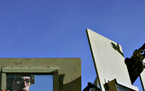 A soldier wearing glasses and combat gear is visible through the armored window port of a military vehicle, likely a HMMWV (Humvee) or similar tactical vehicle. The vehicle is painted in a tan/olive drab color scheme and is equipped with a mounted weapon system on the turret ring. The image is taken from a low angle against a clear blue sky with pine trees visible in the background.