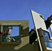A soldier wearing glasses and combat gear is visible through the armored window port of a military vehicle, likely a HMMWV (Humvee) or similar tactical vehicle. The vehicle is painted in a tan/olive drab color scheme and is equipped with a mounted weapon system on the turret ring. The image is taken from a low angle against a clear blue sky with pine trees visible in the background.