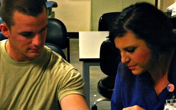 A female medical professional in blue scrubs is administering a blood draw or injection to a young male military service member wearing Army Combat Uniform (ACU). The procedure is taking place at a table in what appears to be a clinical or administrative setting. The service member is watching the procedure being performed on his inner arm.
