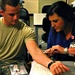 A female medical professional in blue scrubs is administering a blood draw or injection to a young male military service member wearing Army Combat Uniform (ACU). The procedure is taking place at a table in what appears to be a clinical or administrative setting. The service member is watching the procedure being performed on his inner arm.
