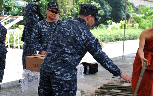 The image shows U.S. Navy personnel in digital camouflage uniforms interacting with a Buddhist monk wearing traditional saffron robes. The setting appears to be an outdoor covered area, likely at a temple or community site, where brooms and bamboo poles are being distributed or organized. This appears to be a community service or humanitarian assistance event.