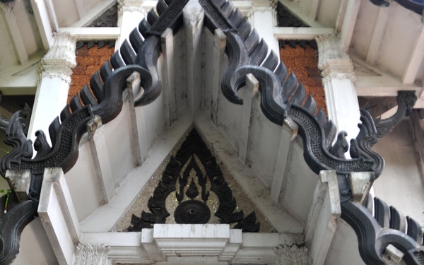 A person wearing blue digital camouflage military uniform and cap stands in the ornate doorway of a Thai Buddhist temple, holding a traditional straw broom. The temple entrance features elaborate white stucco architecture with black naga (serpent) decorations along the roofline and intricate carved details on the columns and pediment. The scene appears to document a community service or humanitarian assistance activity at a Thai religious site.