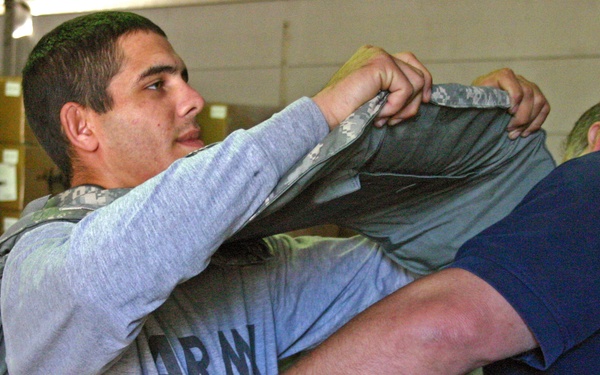 A young male soldier wearing an Army PT shirt is being assisted in putting on or removing a body armor vest (plate carrier) in what appears to be a military supply or equipment room. A second person, visible only partially from behind, is helping with the vest fitting. The background shows stacked cardboard boxes in a warehouse-style environment.