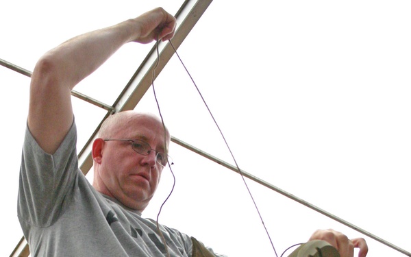 A bald man wearing glasses, a gray t-shirt, dark shorts, and olive green socks is seated on a metal folding chair. He is holding a tan military-style combat boot in one hand while pulling a lace or string upward with his other raised arm. A second person in an Army t-shirt is visible in the background.