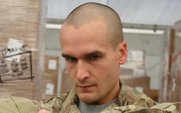 A male soldier wearing MultiCam (OCP) camouflage uniform is extending his fist toward the camera in a posed punching gesture. He is standing in what appears to be a military supply warehouse or logistics facility, surrounded by stacked cardboard boxes. A badge or sticker with a visible American flag design is attached to his chest pocket.