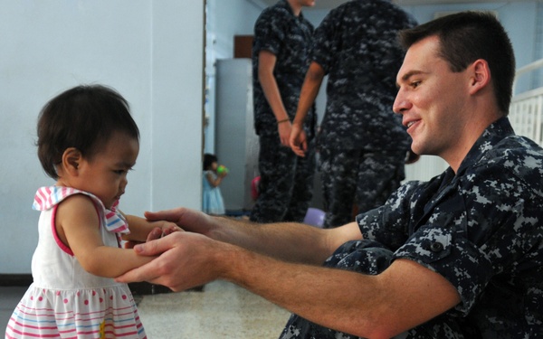 A U.S. Navy sailor in Navy Working Uniform (NWU) sits on the floor of what appears to be a children's care facility or orphanage, interacting warmly with a toddler in a pink and white striped dress. The sailor is smiling and holding the child's hands, while additional sailors in the same uniform are visible in the background. The setting includes infant cribs along the wall, suggesting a nursery or childcare environment.