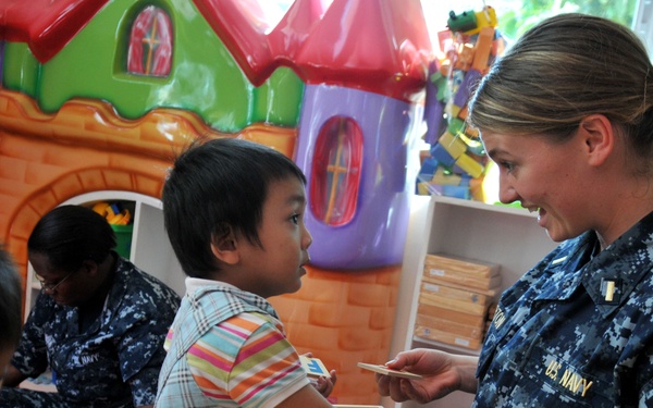 U.S. Navy personnel in blue digital camouflage uniforms are interacting with young children in a colorful classroom or daycare setting. A female sailor is engaging directly with a child using wooden puzzle or flashcard pieces on a table. The scene appears to be a community outreach or humanitarian assistance event.