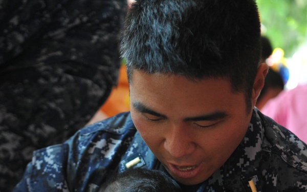 A U.S. Navy service member in digital camouflage uniform is holding a young infant and helping the child interact with colorful plastic toys. The sailor is leaning over the baby attentively, appearing to engage the child in play. The setting appears to be an outdoor or semi-outdoor community or humanitarian event.