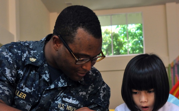 A U.S. Navy sailor in NWU (Navy Working Uniform) digital camouflage is reading a colorful picture book with a young child, likely in a school or childcare setting. The sailor, whose name tape reads 'LLER' (partially visible) and uniform reads 'U.S. NAVY', is pointing at the book while the child looks on attentively. The setting appears to be a community outreach or humanitarian assistance event.