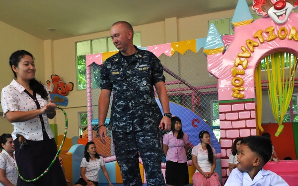 A U.S. Navy servicemember in digital camouflage uniform interacts with children and staff in a colorful indoor play room, likely during a community outreach or humanitarian visit. A young child rides a blue and red toy car while a woman holding a hula hoop converses with the sailor. The setting appears to be a children's recreation facility, possibly in Southeast Asia.