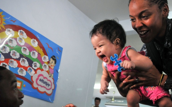 Two U.S. Navy personnel in digital camouflage uniforms interact joyfully with a laughing infant at what appears to be a childcare facility or orphanage. A female sailor holds the baby up toward a male sailor who reaches out with his hands, eliciting a big smile from the child. Several other children and sailors are visible in the background of the room.