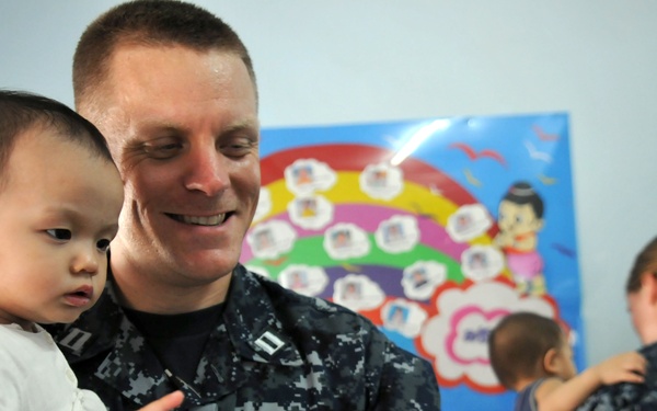 A U.S. Navy officer in digital camouflage uniform is holding a young infant while showing the child something on a smartphone. The scene appears to take place in a childcare facility or orphanage, with a colorful rainbow-themed poster visible on the wall behind them. Additional Navy personnel are visible in the background interacting with other children.
