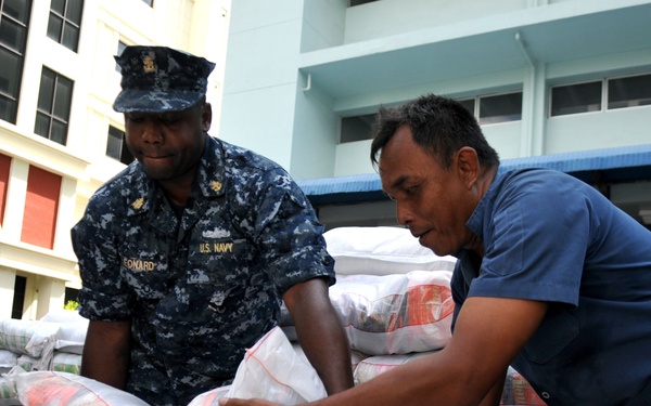 A U.S. Navy sailor, identified by name tape as 'Leonard,' works alongside a civilian man to move large supply bags, likely during a humanitarian aid or disaster relief operation. Additional Navy personnel in the same digital camouflage (NWU Type I) uniforms are visible in the background handling similar bags. The setting appears to be an urban area in a tropical or Southeast Asian location based on the architecture.