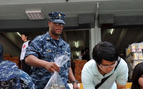 U.S. Navy personnel in blue digital camouflage uniforms are working alongside civilians to sort and distribute relief supplies, likely in a humanitarian aid operation. The scene shows multiple people packing canned goods and other food items into plastic bags near a building stocked with supplies. A Red Cross volunteer is visible in the background, suggesting an organized disaster relief effort.