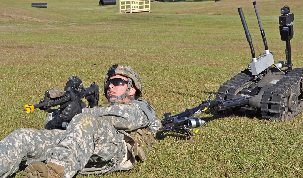 A U.S. Army soldier in ACU (Army Combat Uniform) is lying prone on a grass field alongside a tracked ground robot, likely during a training exercise. The soldier is armed with an M4 carbine fitted with accessories and is wearing a combat helmet and ballistic sunglasses. The scene appears to be a military training range demonstrating human-robot teaming concepts.