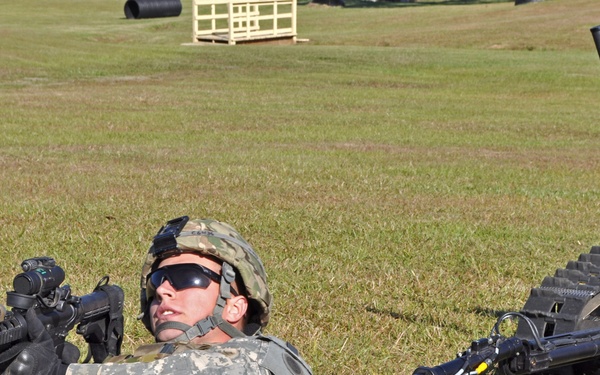 A U.S. Army soldier in ACU (Army Combat Uniform) is lying prone on a grass field alongside a tracked ground robot, likely during a training exercise. The soldier is armed with an M4 carbine fitted with accessories and is wearing a combat helmet and ballistic sunglasses. The scene appears to be a military training range demonstrating human-robot teaming concepts.