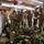 A group of military personnel in desert camouflage uniforms are gathered inside a large warehouse or hangar facility. They appear to be conducting a gear inspection or inventory check, with equipment and supplies spread across the floor in front of them. One individual appears to be holding a clipboard or document, suggesting an administrative or accountability function.