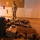 A military service member in camouflage uniform stands behind a large collection of military gear and equipment laid out on a floor inside a building. The equipment appears to be organized for inspection, inventory, or pre-deployment staging. The setting appears to be a military facility or warehouse.