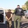 Operation Pipeline Express Drug Arrests An ICE (Immigration and Customs Enforcement) officer is escorting a handcuffed individual toward an open white vehicle door in what appears to be a residential suburban area. The officer is wearing a black tactical vest clearly marked 'POLICE ICE' with yellow lettering on the sleeves. The scene takes place on a paved road during daylight hours with multiple vehicles visible in the background.