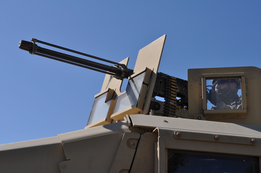 The image shows a close-up, low-angle view of a military vehicle's weapon turret system mounted on what appears to be a tan/desert-colored armored vehicle. A multi-barrel rotary cannon (consistent with a M134 Minigun or similar Gatling-style weapon) is visible along with an ammunition belt feed. A soldier in military gear and eye protection is visible through a side viewport of the vehicle.