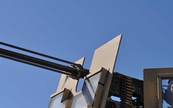 The image shows a close-up, low-angle view of a military vehicle's weapon turret system mounted on what appears to be a tan/desert-colored armored vehicle. A multi-barrel rotary cannon (consistent with a M134 Minigun or similar Gatling-style weapon) is visible along with an ammunition belt feed. A soldier in military gear and eye protection is visible through a side viewport of the vehicle.